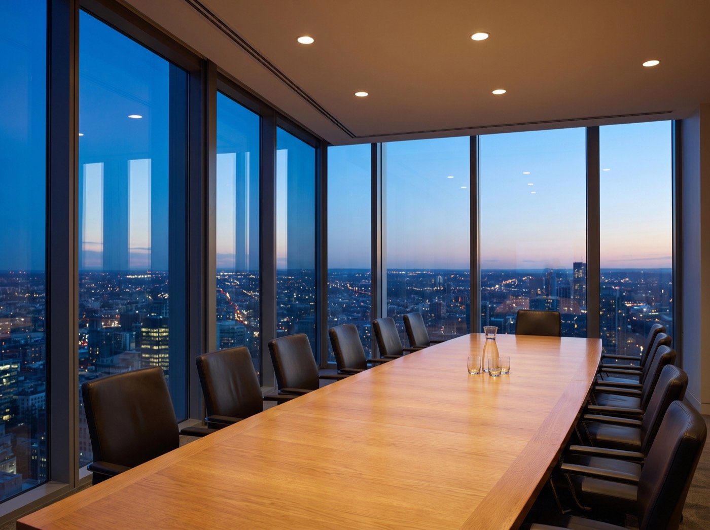 Empty boardroom table beside windows overlooking a city at dusk
